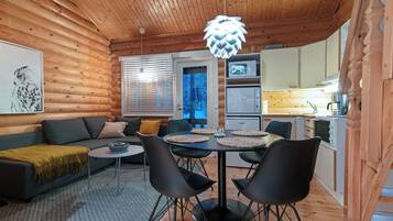 A cozy cabin interior with a wooden ceiling, a dining area with a round table and chairs, a sofa with cushions, and a kitchen with appliances.