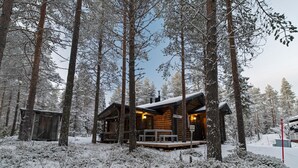 Une cabane en rondins dotée d’un porche et d’une petite terrasse, entourée de grands arbres et d’un sol recouvert de neige.