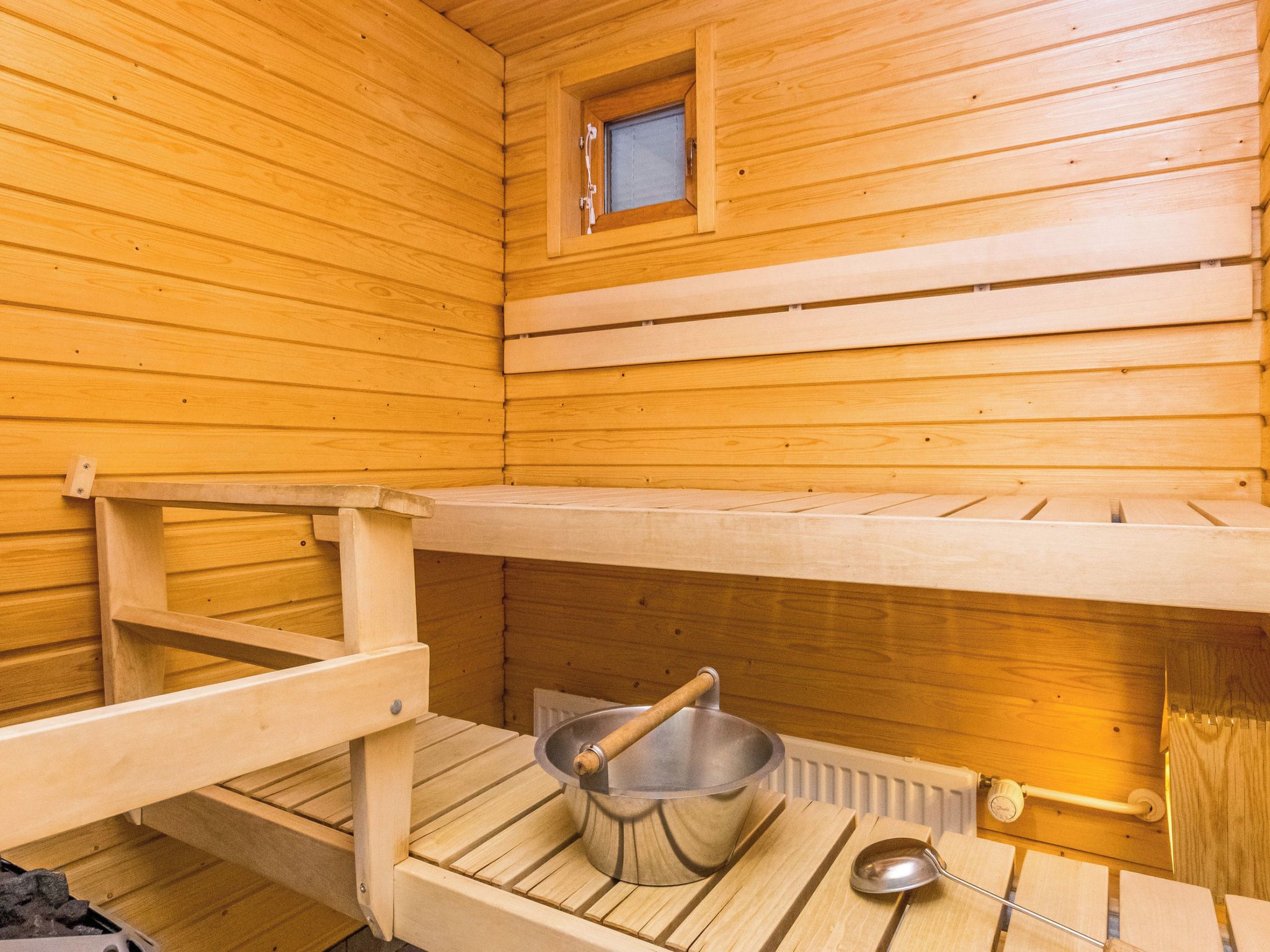 Un sauna avec des bancs en bois, une petite fenêtre et un récipient en métal accompagné d’une louche en bois.