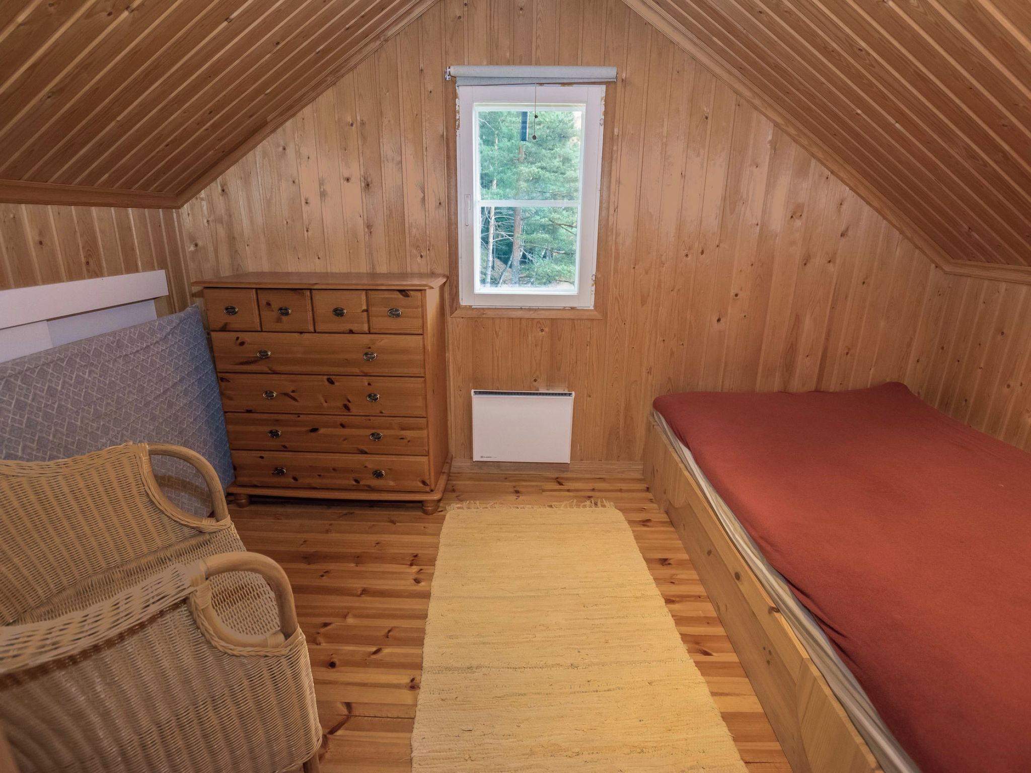 Un pequeño dormitorio en ático con paredes de madera, una ventana, un aparador, una cama y una cesta tejida.