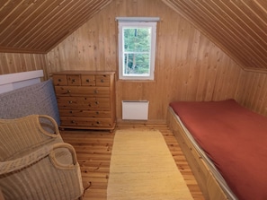 A small attic bedroom with wooden walls, a window, a dresser, a bed, and a woven basket. - Satulinna by Interhome (Kemiönsaari)