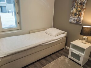 A small, single-bed room with a window, a bedside table with a lamp, and a wall-mounted artwork.