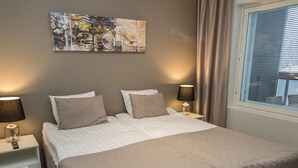 A neatly made bed with a quilted cover, two bedside tables with lamps, a wall-mounted abstract painting, and a window with blinds.