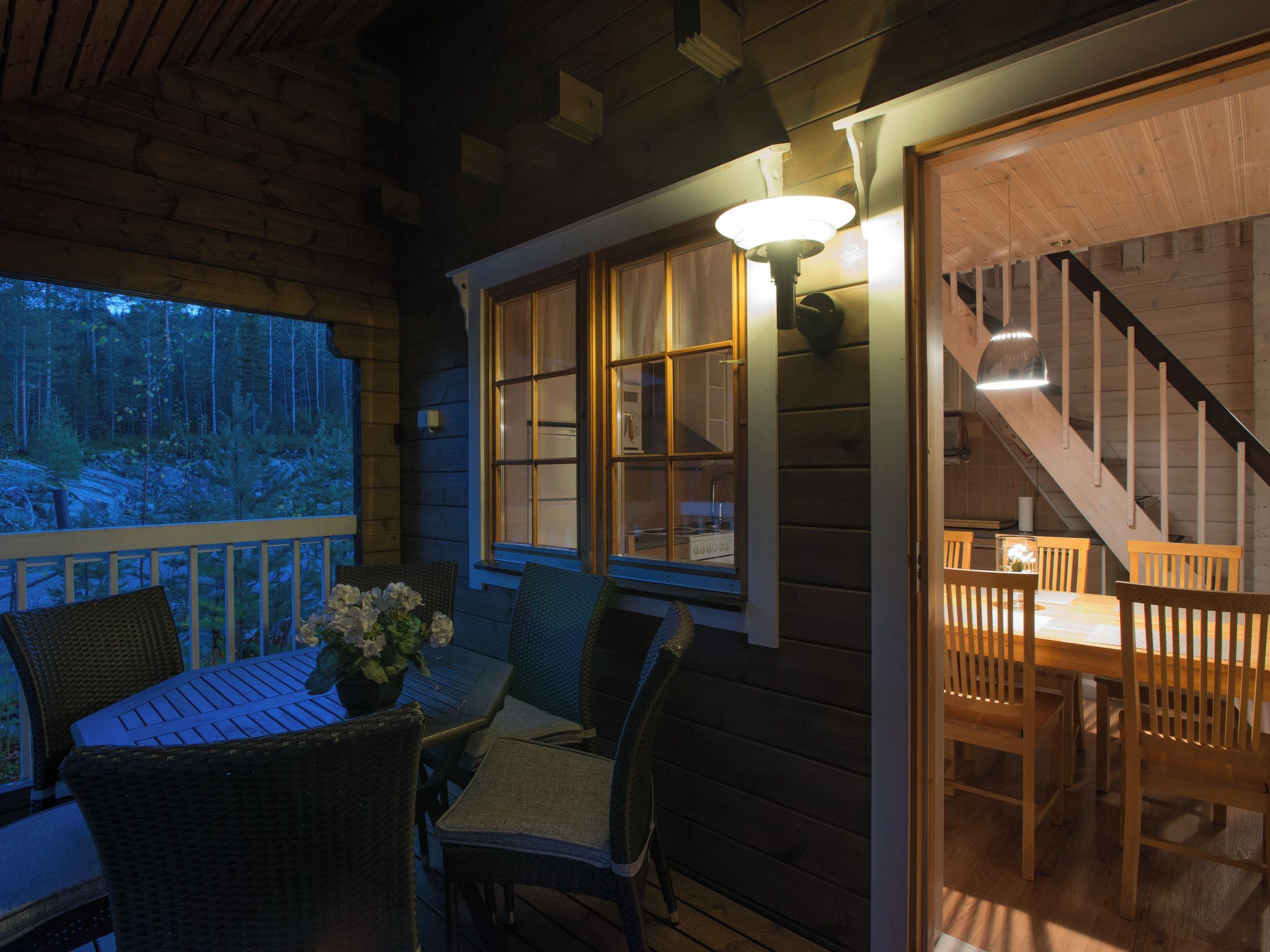 Una terraza de madera con una mesa y sillas, una escalera que sube a un nivel superior, y vista a un bosque a través de una ventana.