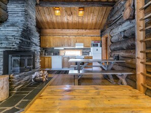 A log cabin interior with a stone fireplace, wooden cabinets, a kitchen area with appliances, and a picnic table.