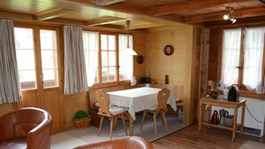 A cozy cabin interior with a wooden dining table, two leather chairs, a small kitchen area, and large windows with curtains. - Marmotte, Chalet by Interhome (Lauenen b. Gstaad)