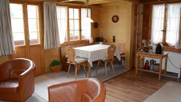 Ein gemütliches Blockhaus-Interieur mit einem Holzespeisetisch, zwei Ledersesseln, einer kleinen Küchenzeile und großen Fenstern mit Vorhängen.