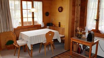 Ein gemütliches Blockhaus-Zimmer mit Holzvertäfelung, einem Esstisch mit Stühlen, einer kleinen Küchenzeile inklusive Kaffeemaschine und einem Fenster mit Vorhängen.