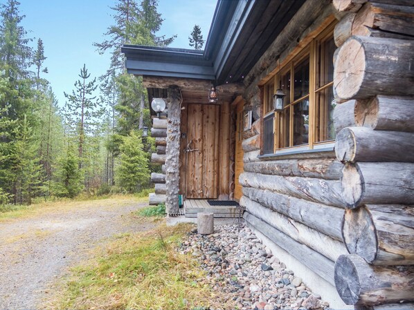 Una cabaña de troncos con puerta y ventanas de madera, rodeada por un camino de grava y un bosque.