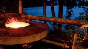 Une table en bois sur laquelle brûle un feu, entourée de chaises, le tout se dressant sur un fond de forêt et au bord d’une étendue d’eau.