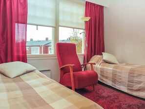 A room with two beds, a red chair, a lamp, and a window with a view of buildings.