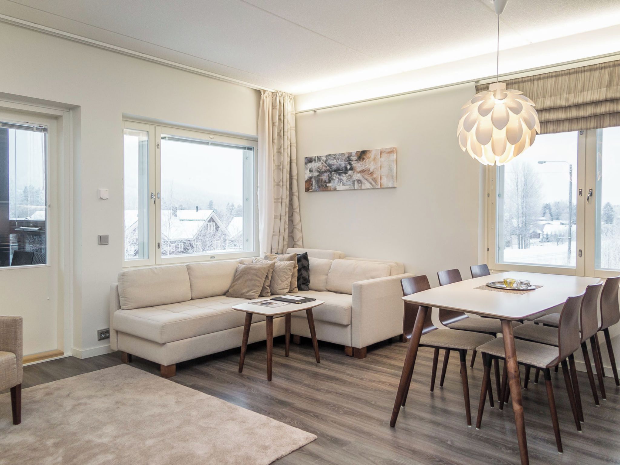 A modern living room with a sofa, coffee table, dining table, and chairs. There are large windows with curtains, a painting on the wall, and a pendant light.