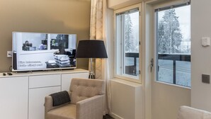 Una camera d'albergo con una televisione che mostra una scena, una lampada, una sedia e una finestra vista alberi e un paesaggio innevato.