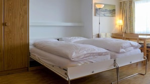 A single bed with a metal frame, a wooden wardrobe, a desk with a chair, and a lamp.