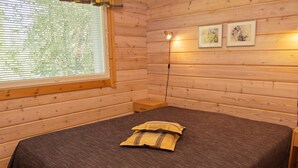 A wooden cabin room with a bed, a window with blinds, a bedside table, and two framed pictures on the wall.