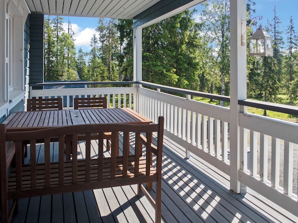 A covered outdoor deck with a wooden table and benches, surrounded by white railings and a view of trees and a distant building.