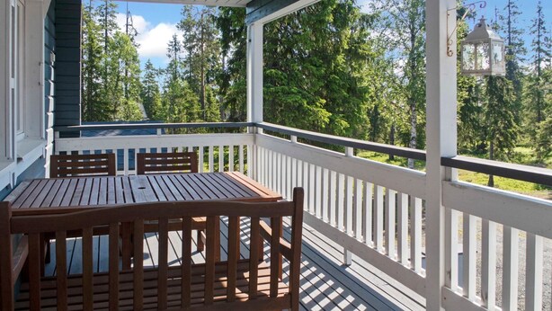 A wooden deck with a table and chairs, surrounded by white railings, overlooking a forested area.