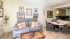 Ein Wohnzimmer mit grauem Sofa, einem Couchtisch mit Pflanze, einem Essbereich mit Korbstühlen und einer Küche im Hintergrund.