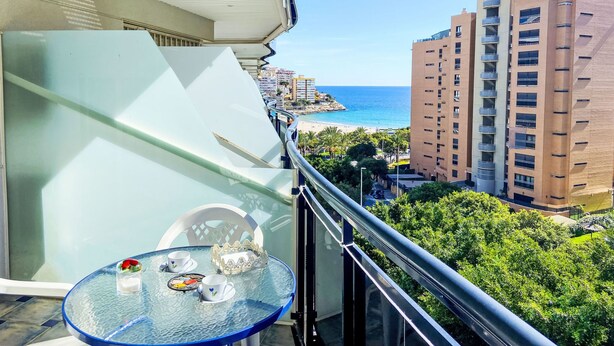 Ein Balkon mit Glastisch, einem Stuhl und einem dekorativen Gegenstand. Blick auf einen Strand und mehrere Gebäude.