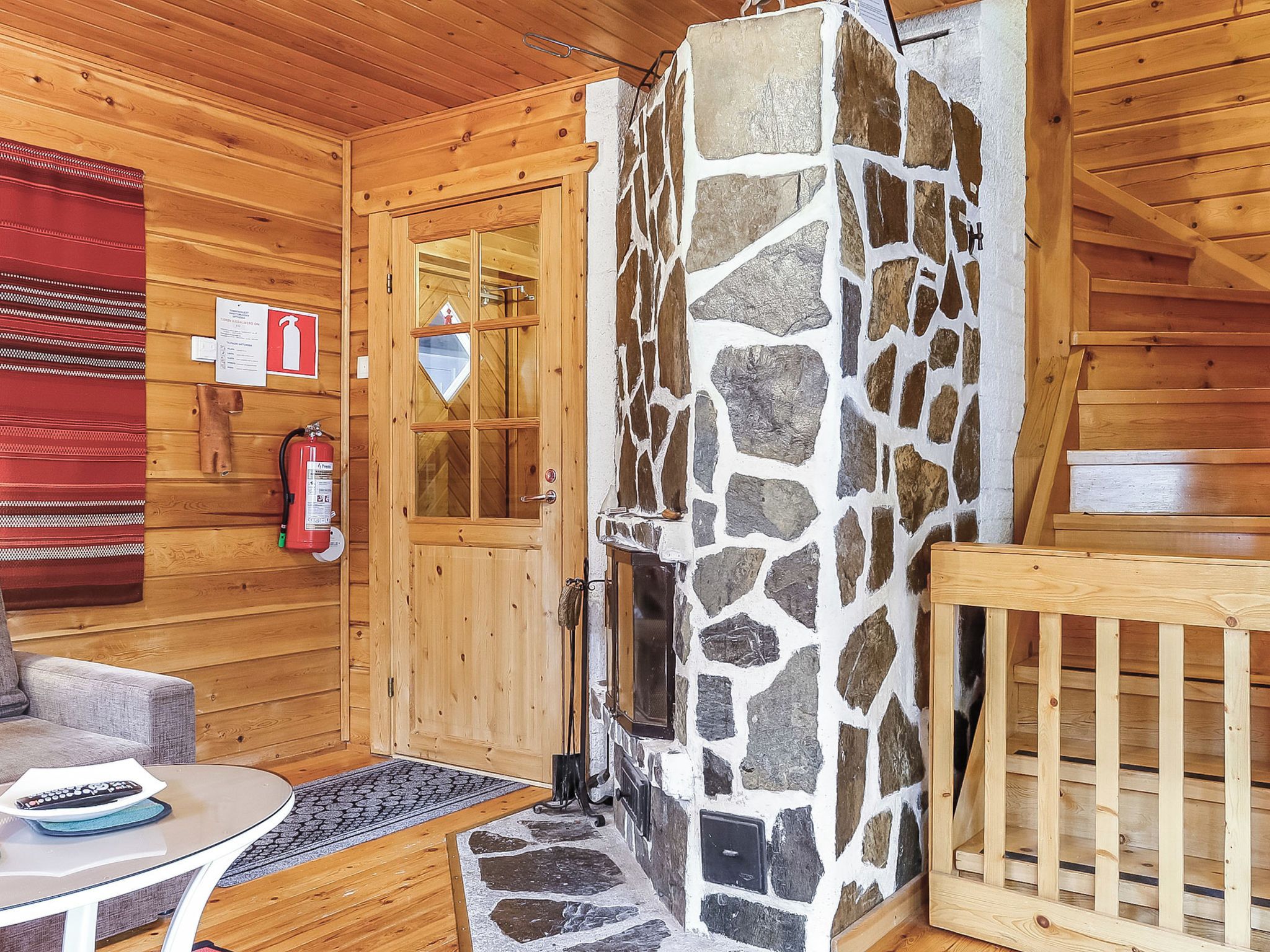 A wooden cabin interior with a stone fireplace, a wooden door, a fire extinguisher, and a red blanket on the wall.