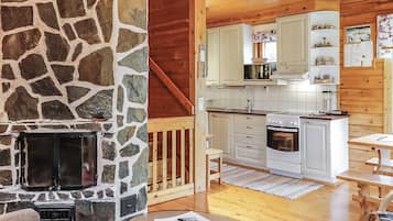A cozy cabin interior with a stone fireplace, wooden flooring, a kitchen with white cabinets, and a dining area with a table.