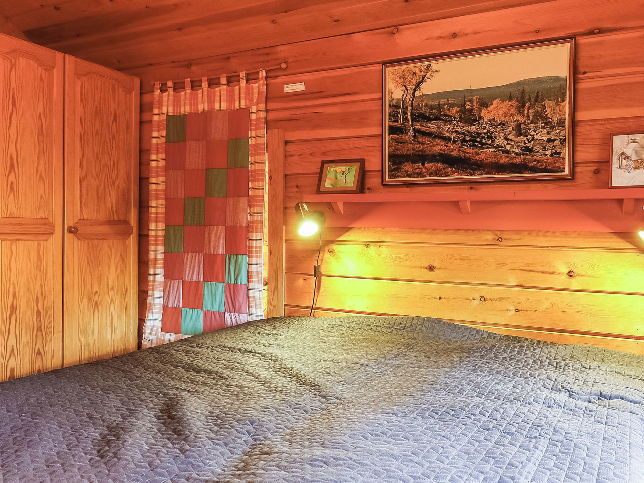 Ein Zimmer in einer Holzhütte mit einem Bett, einer gesteppten Matratze, einem karierten Vorhang und einem gerahmten Bild an der Wand.