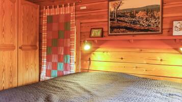 Ein Zimmer in einer Holzhütte mit einem Bett, einer gesteppten Matratze, einem karierten Vorhang und einem gerahmten Bild an der Wand.