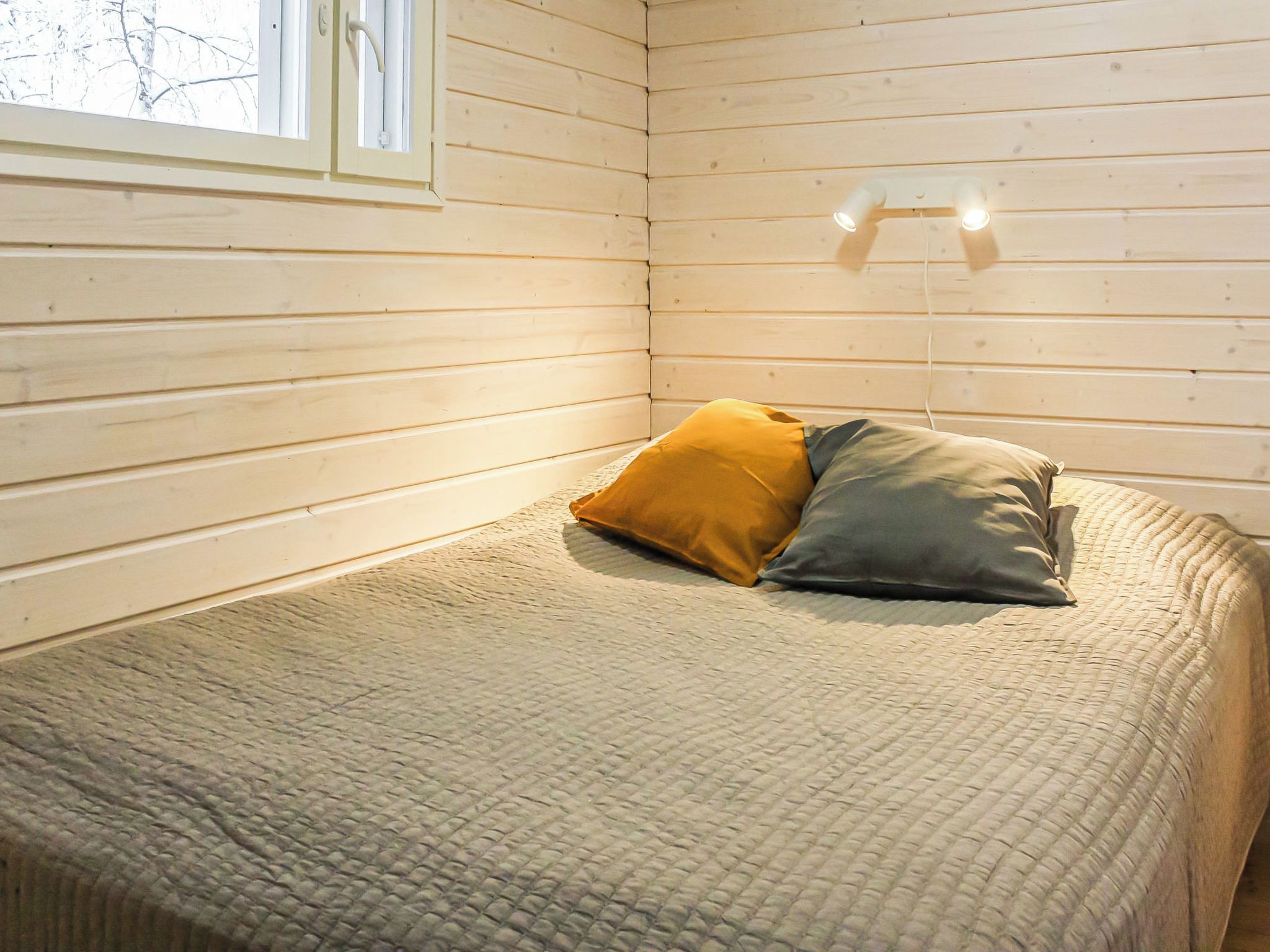 A room with wooden walls, a bed with a textured blanket, two pillows, and a window with blinds.