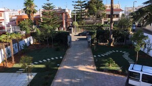 A residential area with a paved pathway leading to a gated entrance, surrounded by greenery and palm trees.