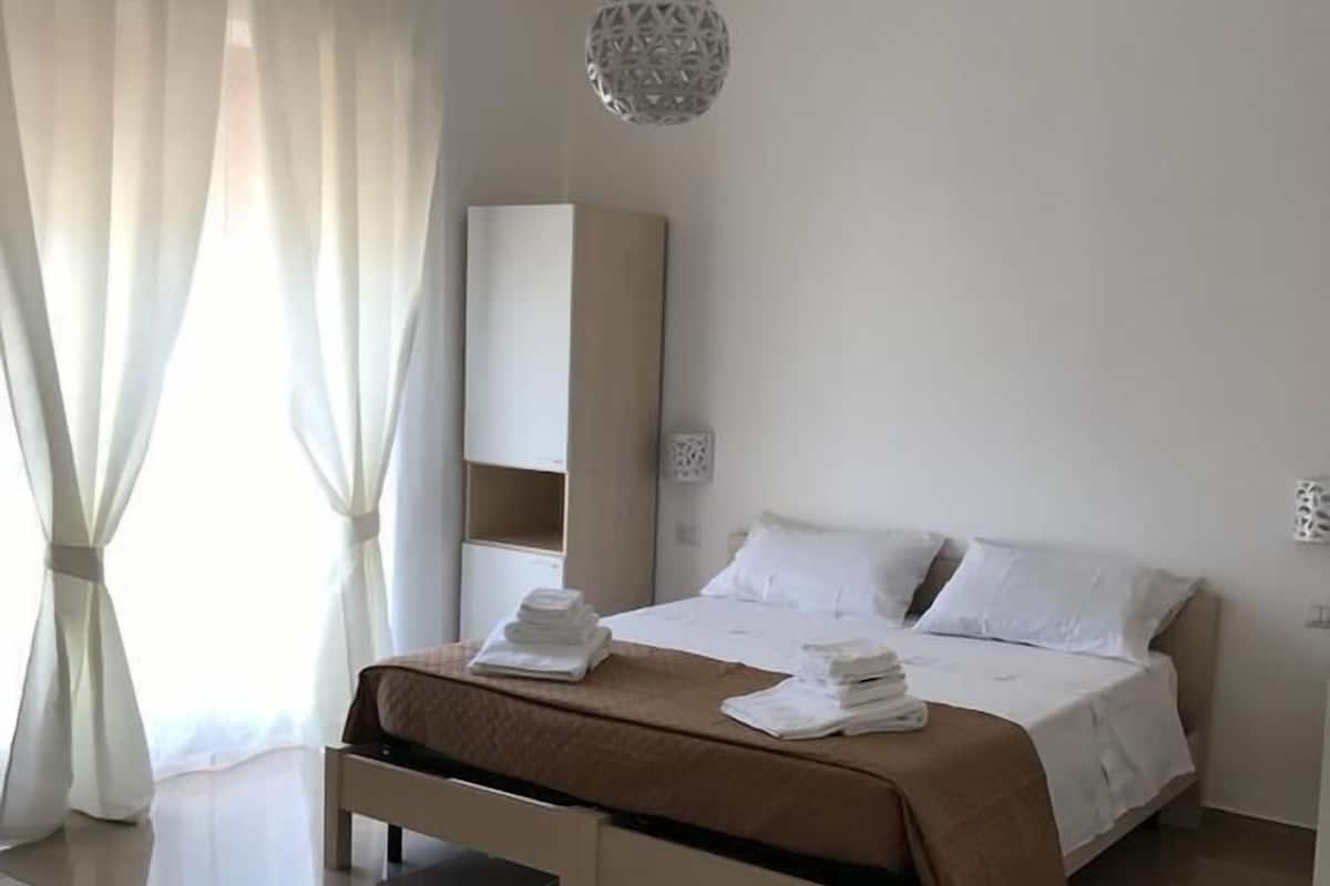 A hotel room with a bed, a bedside table, a hanging light fixture, and a large window with curtains.