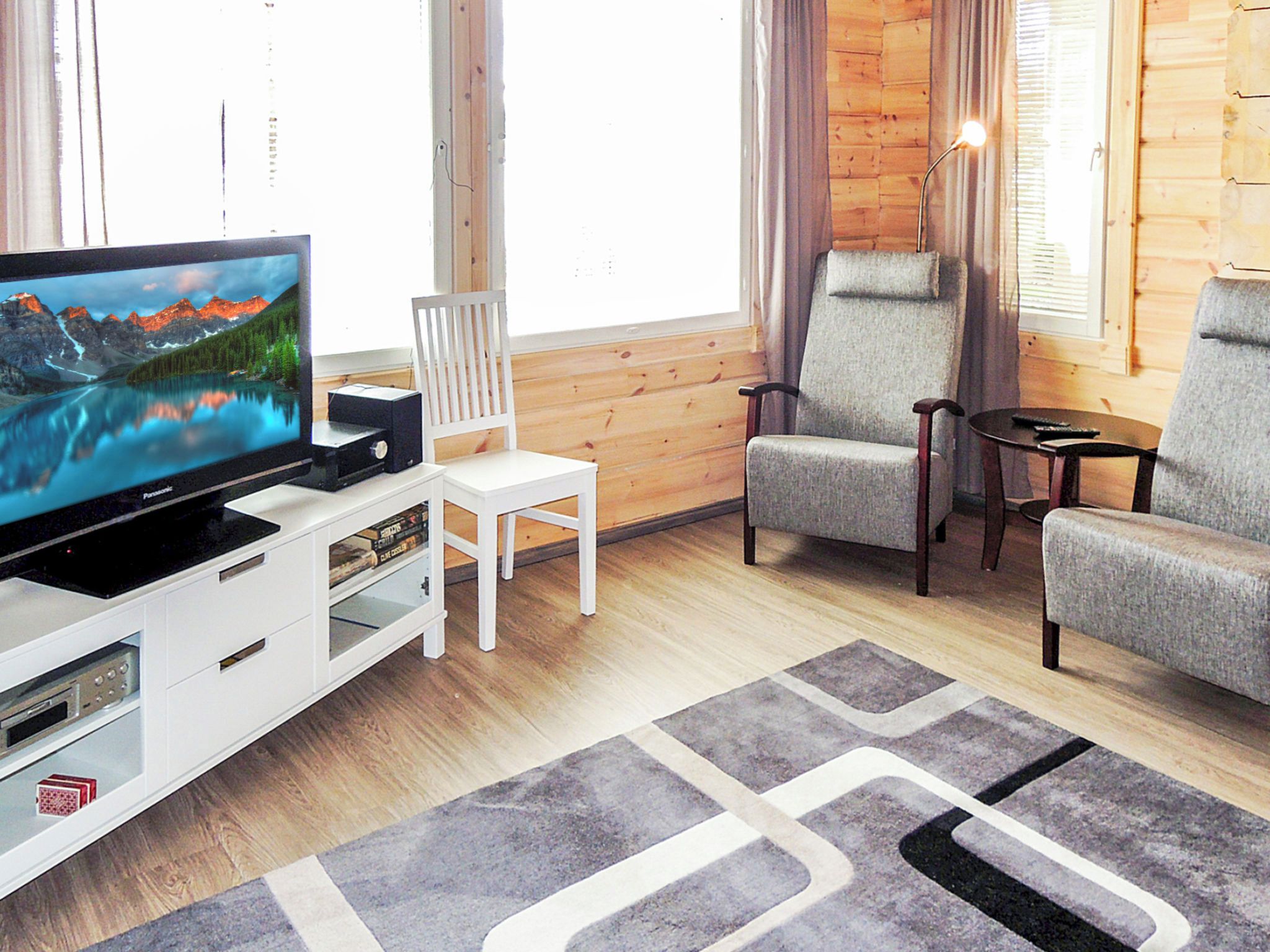 A living room with a flat-screen TV, a white entertainment unit, two grey armchairs, a white chair, a small table, and a patterned rug.