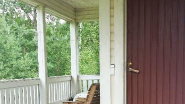 A covered porch with a wooden bench and a chair, a red door, and a welcome sign.