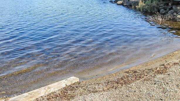 Un lago tranquilo con aguas cristalinas, una orilla rocosa y un área boscosa en la distancia.