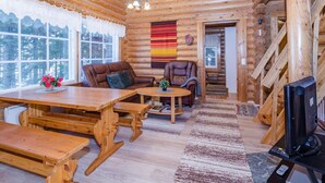 Interior acogedor de una cabaña de troncos con una mesa de comedor de madera, dos sofás de cuero, un televisor y un ventanal con vistas a un paisaje nevado.