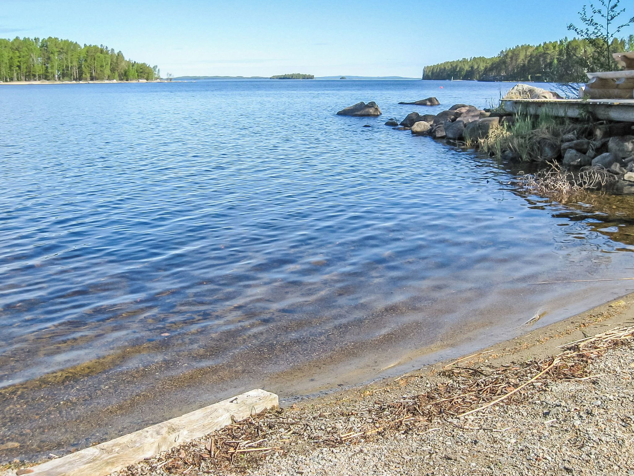 A calm lake with clear water, surrounded by a rocky shoreline and dense forest.