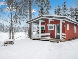 Punainen mökki, jossa on valkoinen kuisti ja ovi, ja sen ympärillä on lumipeitteisiä puita.