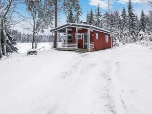 Una cabaña nevada con un porche, rodeada de árboles y un paisaje cubierto de nieve.