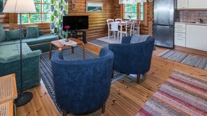 Un acogedor interior de cabaña con un sofá, dos sillones, una mesa de comedor, un televisor y una zona de cocina.