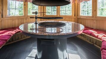 A round table with a grill in the center, surrounded by wooden walls and windows with checkered curtains.