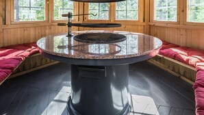 Una mesa de piedra circular con una rejilla metálica en una cabaña de madera con cortinas escocesas y bancos de madera.