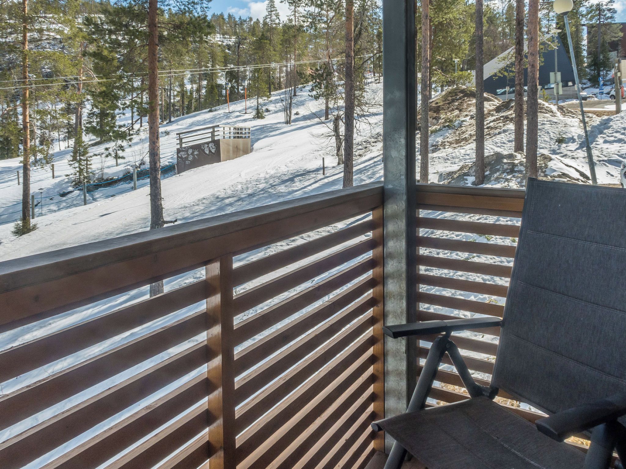 Una terraza de madera con una silla, con vista a un paisaje nevado con árboles y un telesilla.