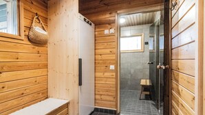 Interior de una cabaña de madera con un banco, un refrigerador blanco, un box de ducha de vidrio y una puerta de madera.