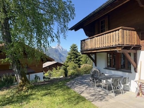 A wooden chalet with a balcony, a dining area with chairs and a table, and a view of mountains in the background.