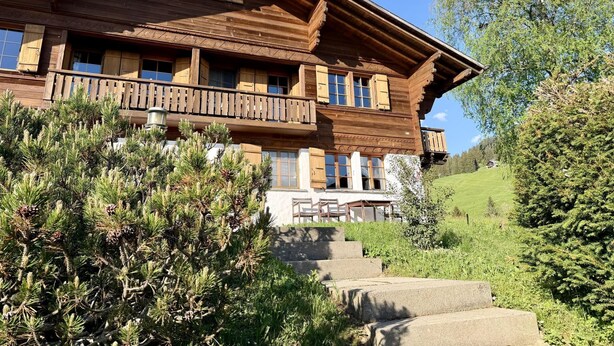 A wooden chalet with a balcony, surrounded by greenery and a clear sky.