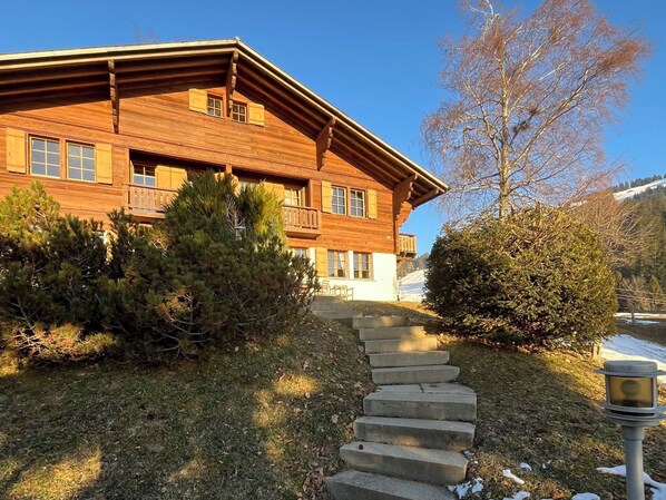 A wooden lodge with a staircase leading to the entrance, surrounded by trees and a clear sky.