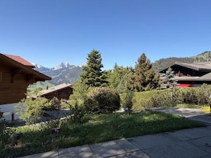 A wooden cabin with a mountain backdrop, surrounded by greenery and a clear sky.
