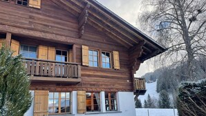 A wooden chalet with a balcony, a covered outdoor seating area, and a snowy landscape in the background.