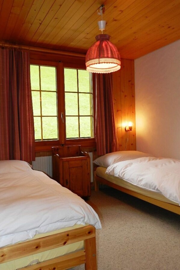 A room with two wooden beds, a wooden desk, a window with curtains, and a hanging lamp. - Hornflue (Baumann) by Interhome (Saanen)