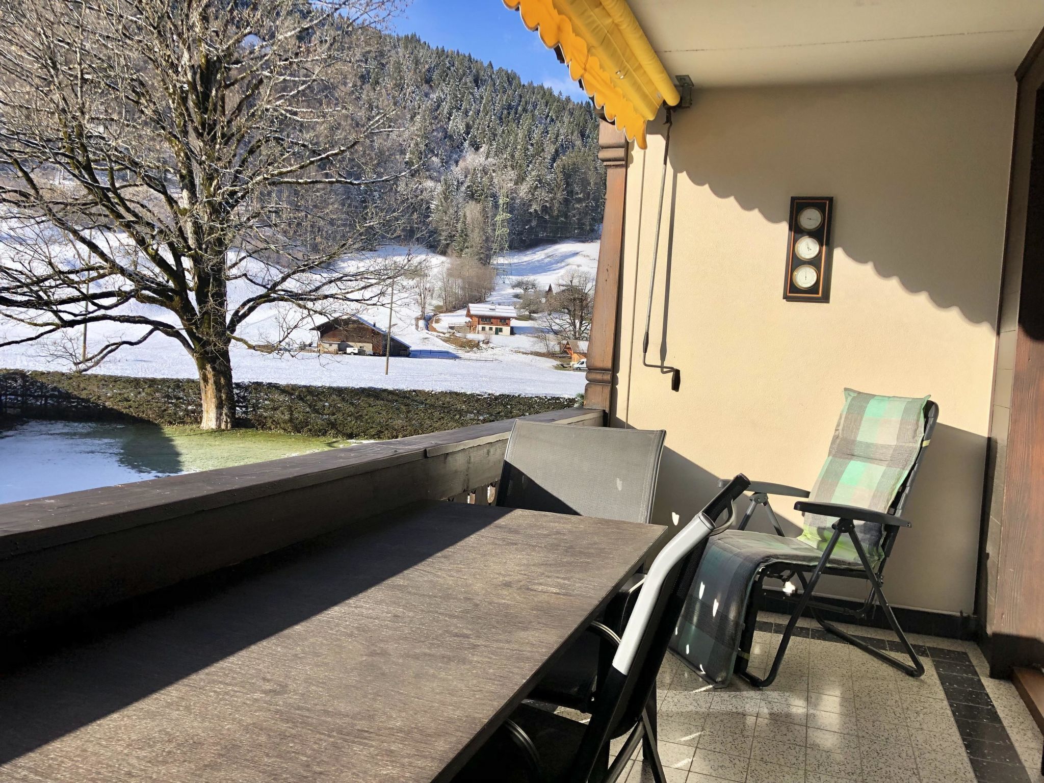 Un balcón con una mesa y sillas, vista a un paisaje nevado con árboles y casas, y un reloj en la pared.