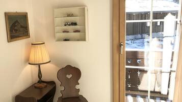 Ein Zimmer mit Holzstuhl, Lampe, einer Kommode mit Modellautos und einer Tür, die auf einen Balkon mit Blick auf eine verschneite Landschaft führt.